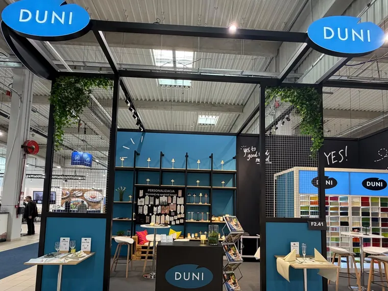Front stoiska: pergola z dwoma świecącymi owalnymi logo Duni, niebieska ściana z regałem i czarna lada z logo
