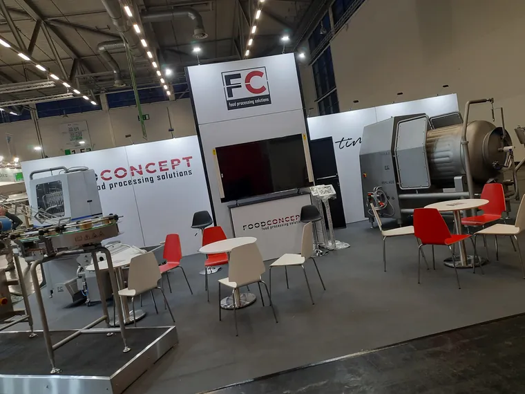 Stoisko z białymi ścianami, wieżą z logo FC nad zapleczem, ekranem i maszynami do przetwórstwa na froncie