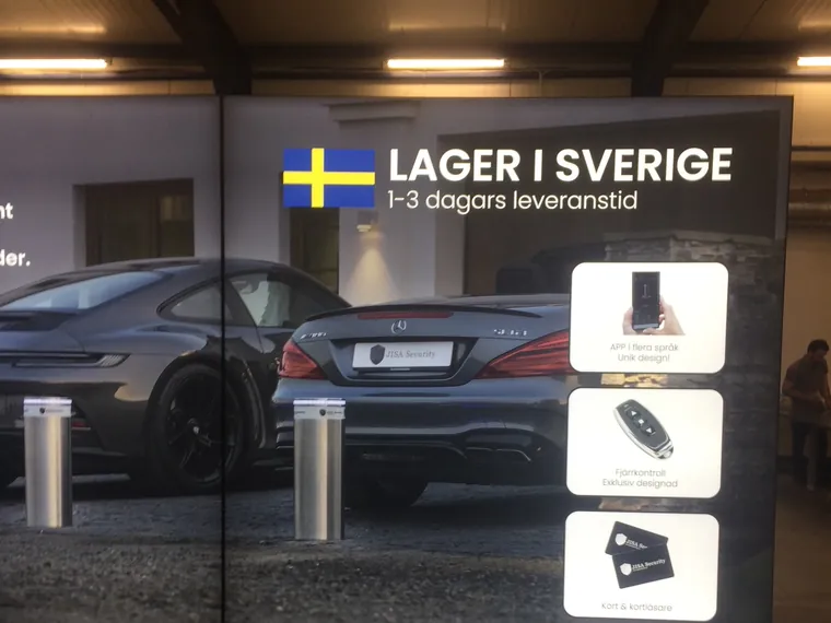 Detal ściany: hasło „Lager i Sverige” z flagą Szwecji, słupki przed samochodem i białe pola ze zdjęciami pilota, aplikacji i kart