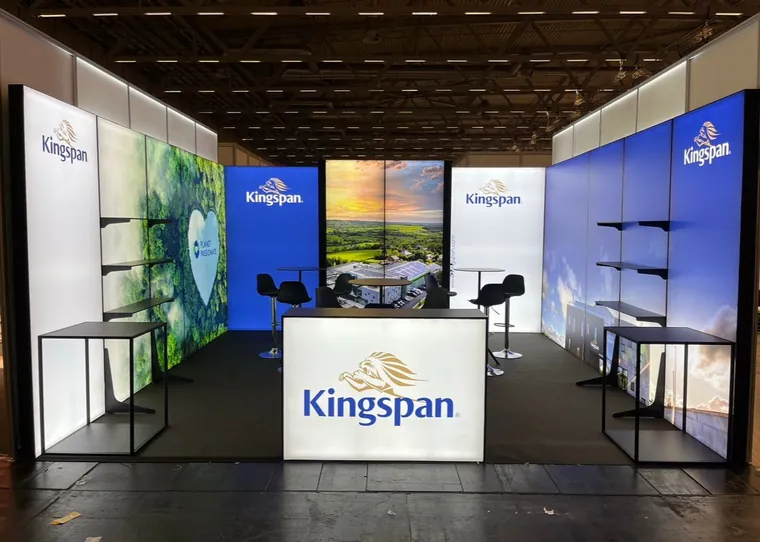 Stoisko z podświetlanymi ścianami z logo Kingspan, zaplecze na środku, półki na ścianach bocznych, stoliki barowe, lada z logo i dwa podesty na froncie